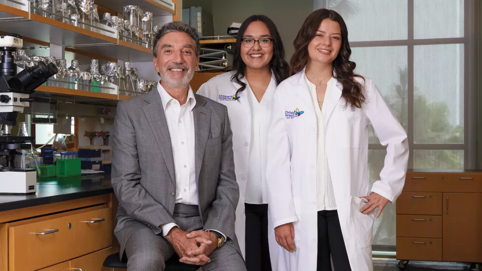 Chuck Lorre, a man with light skin and grey hair, wearing a white shirt and grey suit sits with two female researchers wearing white lab coats in a laboratory setting