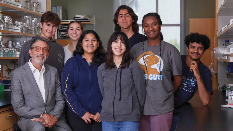 Chuck Lorre, a man with light skin and grey hair, wearing a white shirt and grey suit sits with seven casually dressed high school students in a laboratory setting