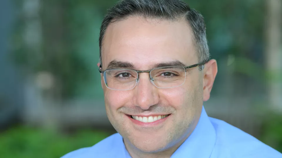 Headshot of smiling man with light skin tone and greying hair wearing eyeglasses, a blue dress shirt and plaid tie against a blurred outdoor background