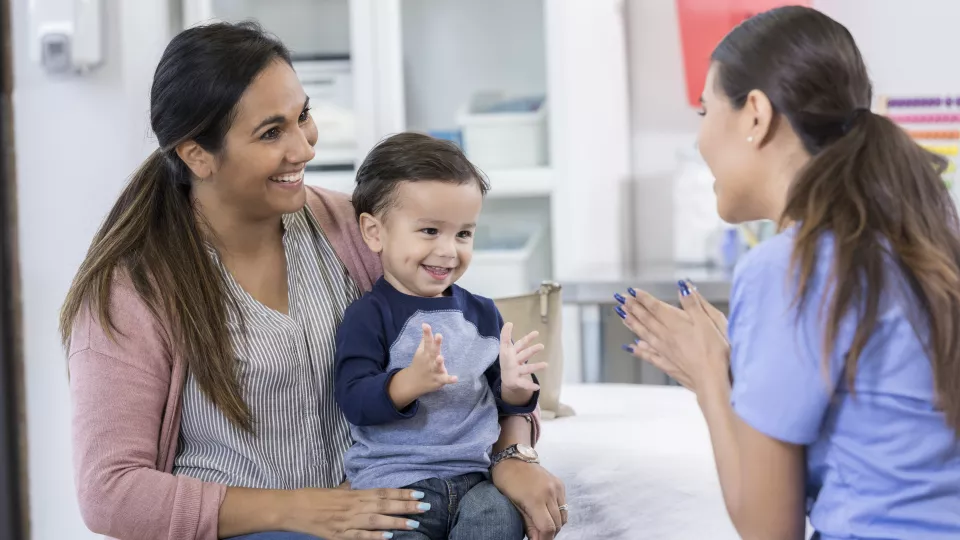 A little boy sits on his mom’s lap across from a nurse in blue scrubs. He is smiling at the nurse and clapping his hands.