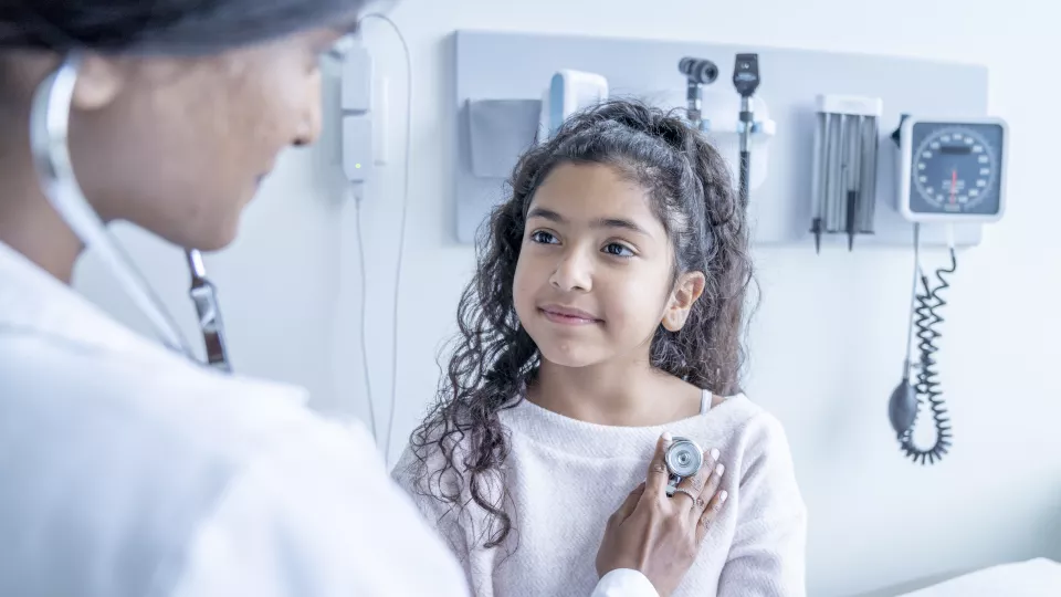 A female doctor with medium skin tone and dark hair listens to her young patient's heartbeat as she sits up on an exam table.