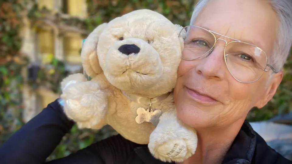 Actress Jamie Lee Curtis wearing gold framed eyeglasses poses with a stuffed dog