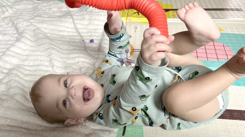 Male infant with light skin tone wearing a light green onesie with car pattern lies on their back and sticks out their tongue