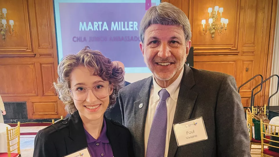 Smiling young woman with light skin tone stands with a smiling adult male with light skin tone and greying hair wearing a grey suit