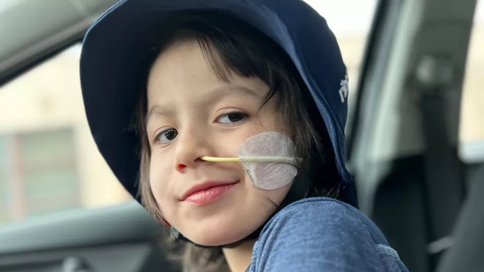 Smiling young boy with medium skin tone, dark hair and a feed tube sits in the passenger seat of a car wearing a blue bucket hat and blue t-shirt