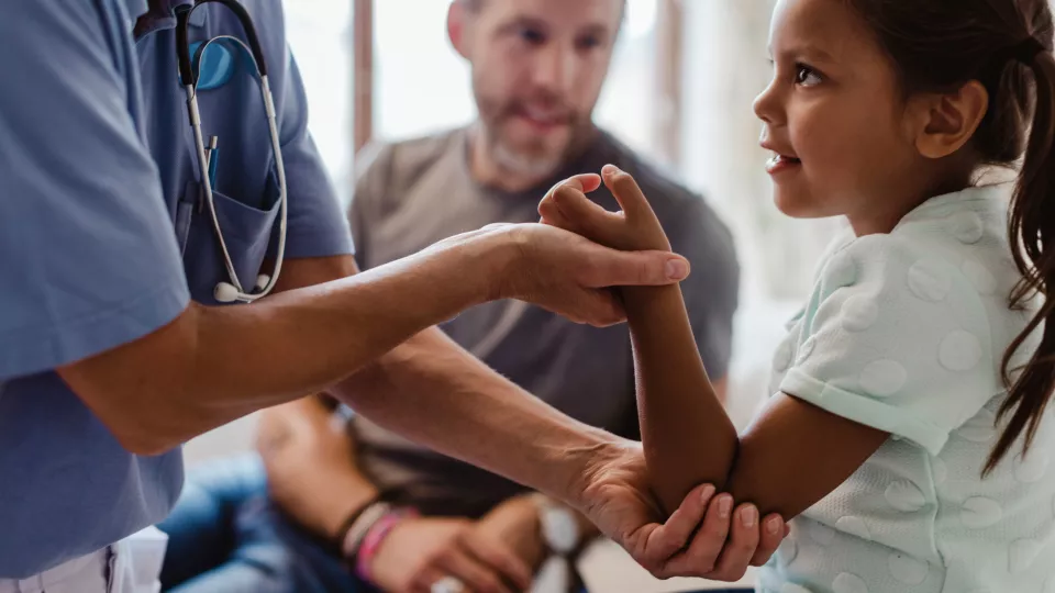 The arms of a nurse wearing blue scrubs are seen checking the arm of a girl with medium skin tone. The girl’s father looks on.