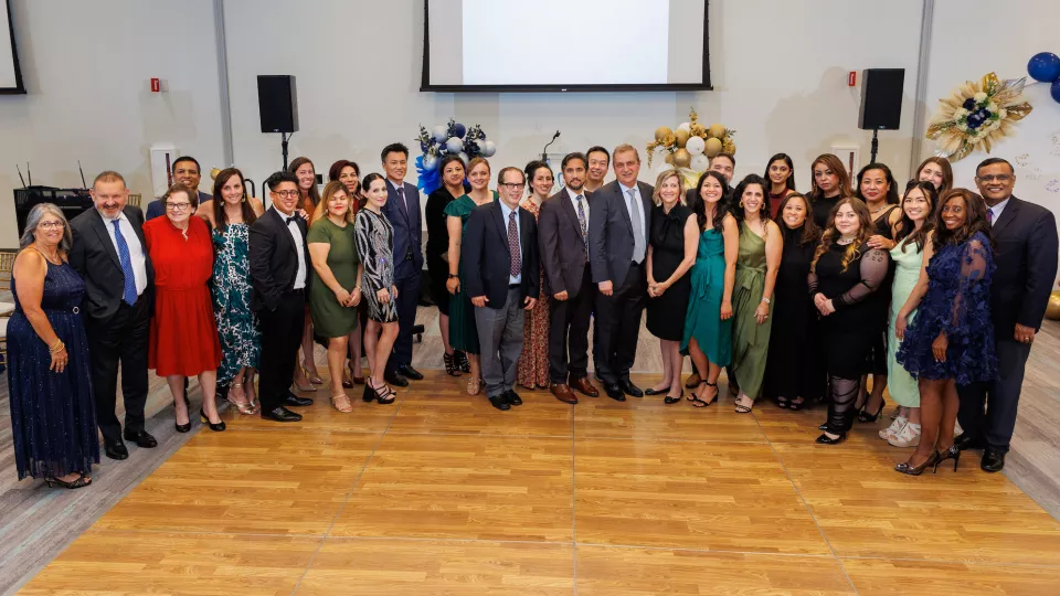 A group of more than 30 adults dressed in suits and gala attire pose in a semi-circle in front of a large projector screen