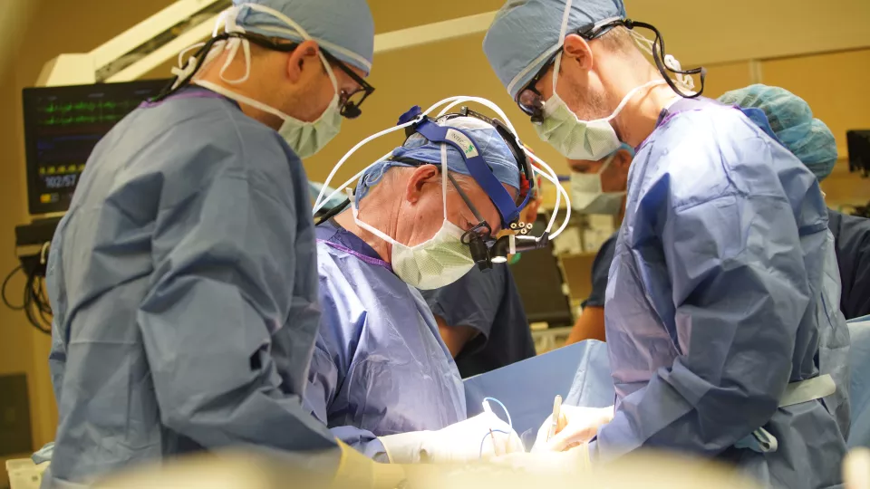 Three male surgeons with light skin tone and wearing surgical gowns, surgical masks and surgical eye loupes perform a surgical procedure on a patient in a hospital operating room