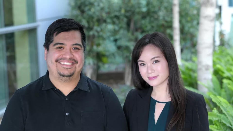 Smiling man with medium skin tone, dark hair and a goatee wearing a black polo shirt stands next to smiling woman with light skin tone and long dark hair wearing a green top under a dark suit jacket against a blurred outdoor background