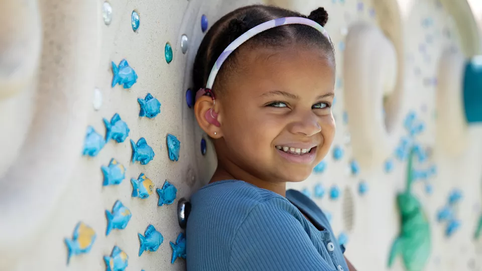 Young girl with dark skin tone and short dark hair wearing a blue sweater smiles as they lean, arms crossed, against a cream-colored decorative wall adorned with molds of brightly colored aquatic animals