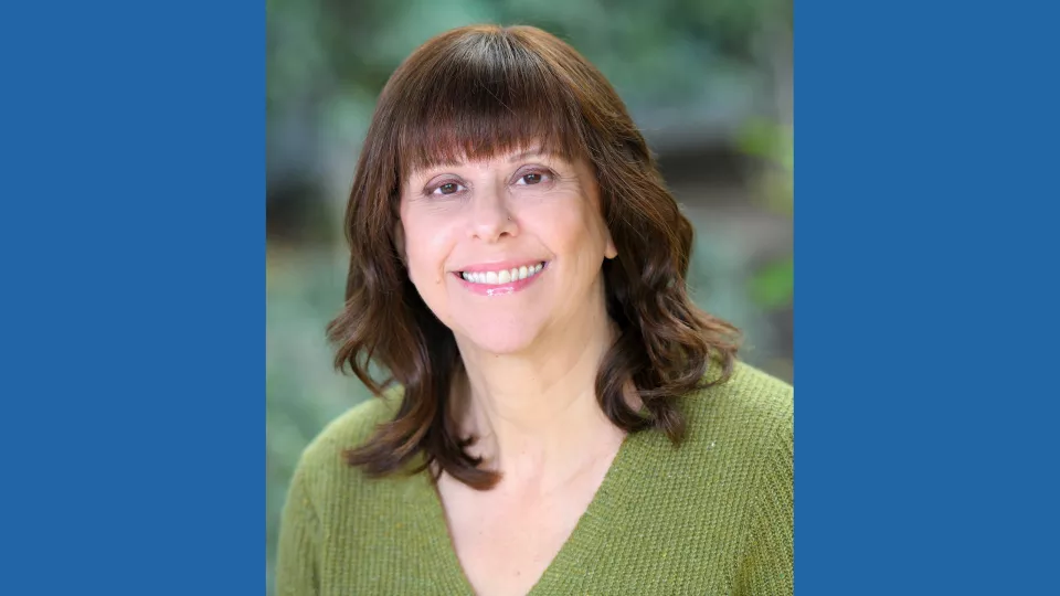 Headshot of a smiling woman with light skin tone and shoulder-length brown hair wearing a v-neck green sweater against a blurred outdoor background