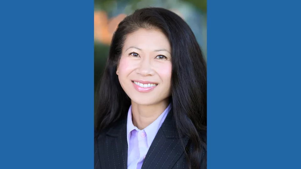 Headshot of a smiling woman with light skin tone and straight dark hair wearing a purple blouse under a dark suit jacket against a blurred outdoor backgroun
