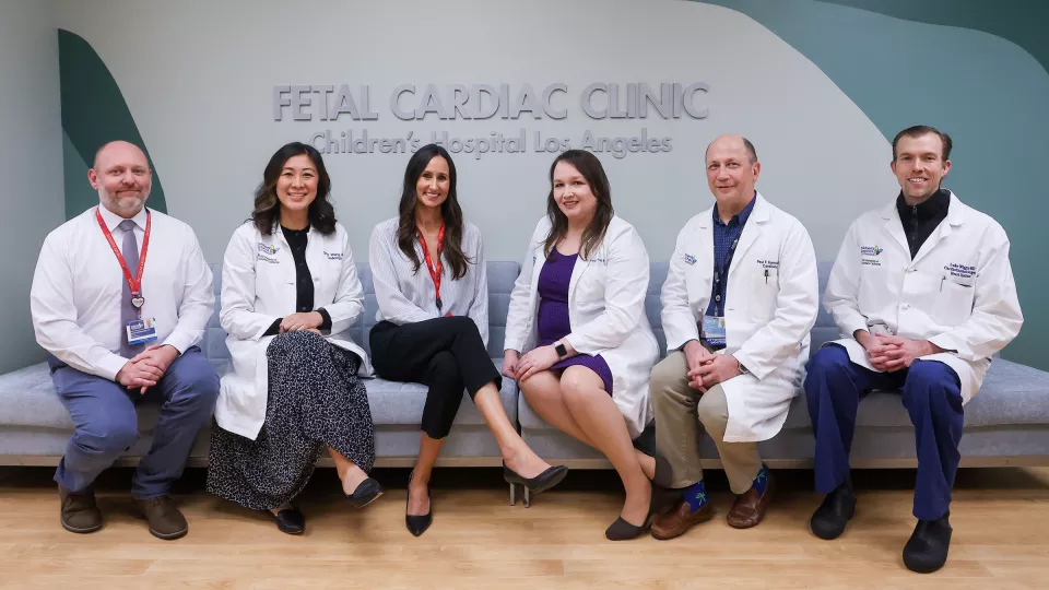 Six health care workers with light skin tone sit on a low grey couch behind a sign that reads Fetal Cardiac Clinic