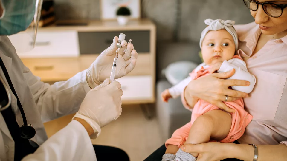 A healthcare worker wearing latex gloves, surgical mask and face shield holds a small vial and syringe and prepares to vaccinate an infant with medium skin tone being held in her mother's arms