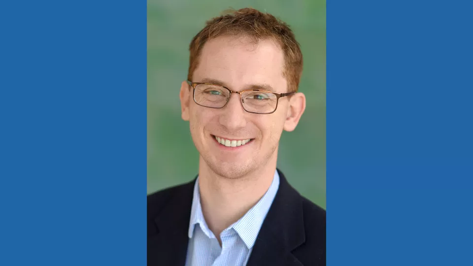 Headshot of a smiling man with light skin tone and auburn hair wearing rectangular eyeglasses and an open-collar light blue dress shirt under a dark suit jacket against a blurred outdoor background