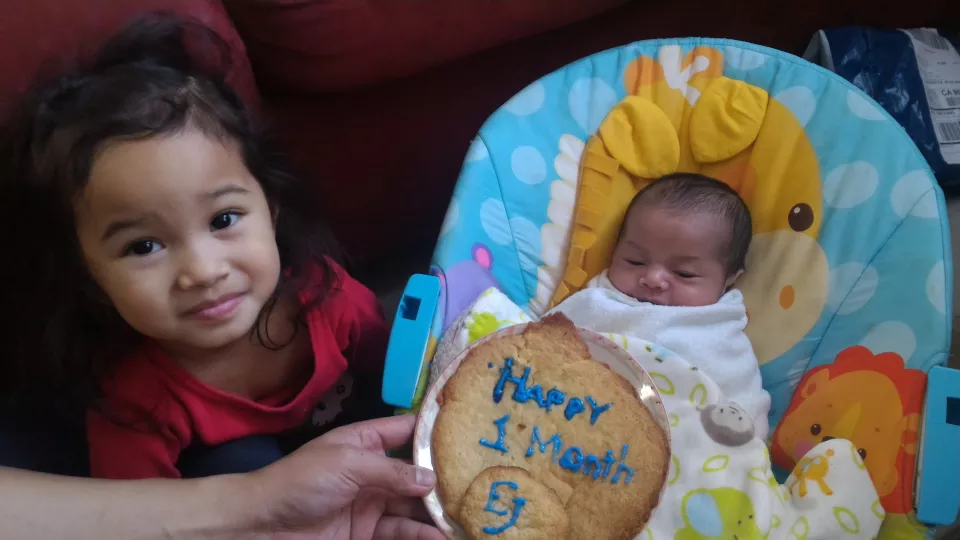 A medium skin toned toddler sits next to a 1-month-old baby swaddled in blankets. In front of them a hand holds a cookie with “Happy 1 Month EJ” written in icing.
