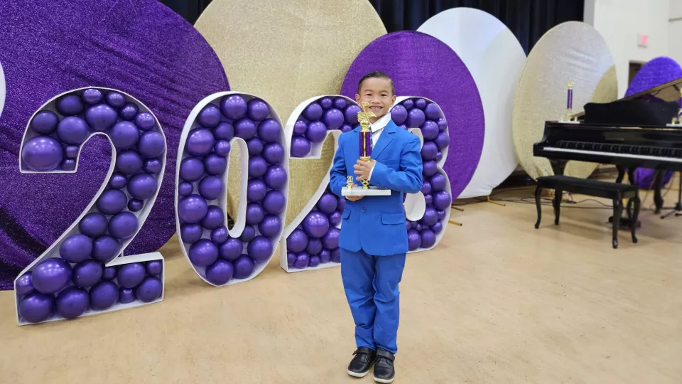A medium skin-toned boy wearing a blue suit and holding a trophy smiles at the camera. Behind him are a piano and large purple decorations spelling out 2023 in balloons.