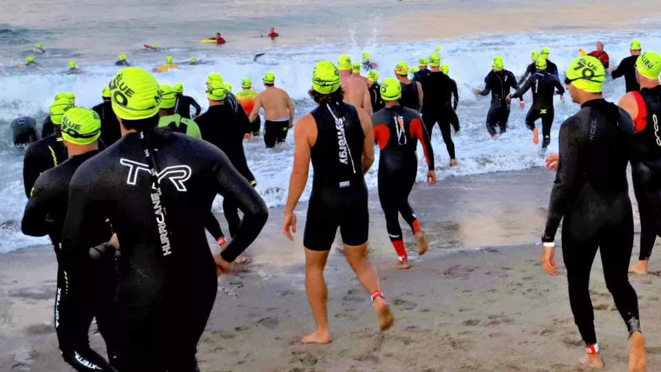 About 50 athletes wearing black wetsuits and bright yellow swim caps head down the beach into the ocean waves beneath a brightening sky in Malibu