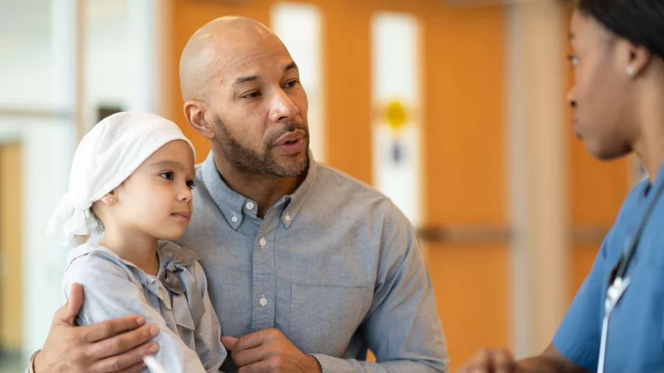 A young girl with a light-colored headscarf and her dad talk with a nurse in blue scrubs. The dad has his arm around his daughter.