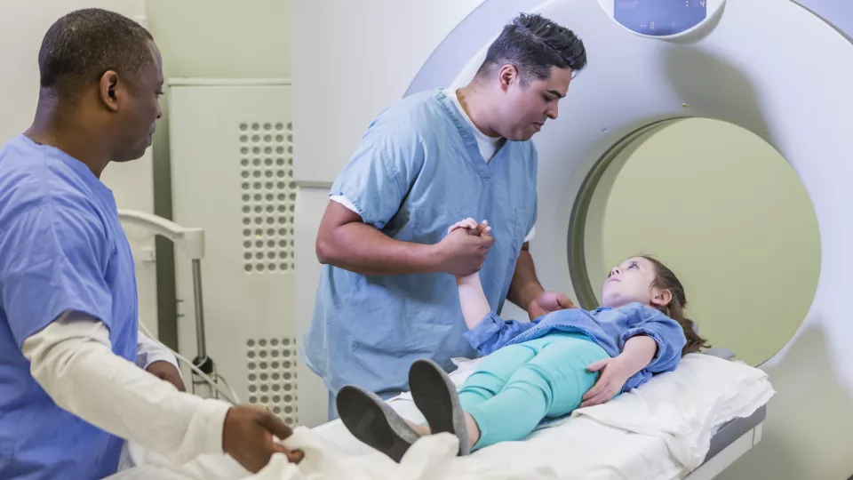 Two health care providers comfort a young girl with light skin tone as she gets ready for a CT scan.