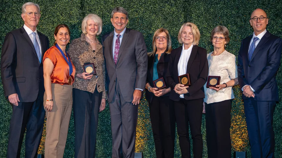 Five women and three men in business attire stand in a row against a green background