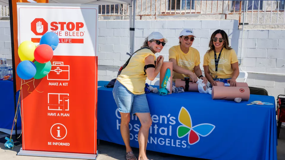 Three women with light skin and yellow shirts standing around table draped in blue linen with logo