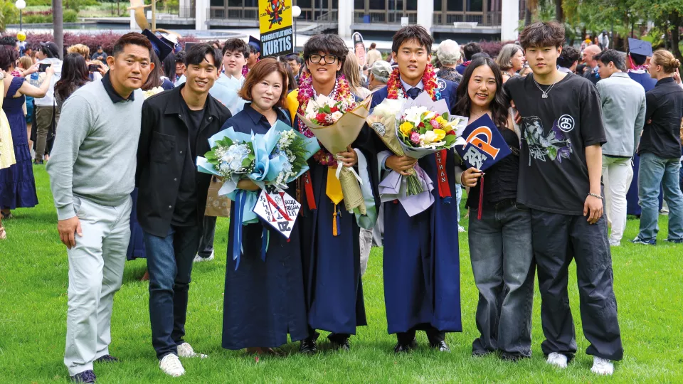 A family of seven, five males and two females all with light skin tone standing outside at a graduation ceremony with two male graduates and an older woman each holding a bouquet of flowers.