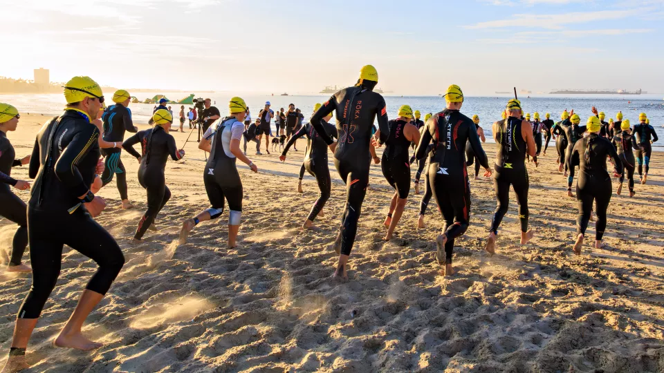 Athletes wearing black wetsuits and bright yellow swim caps run down the beach toward the ocean