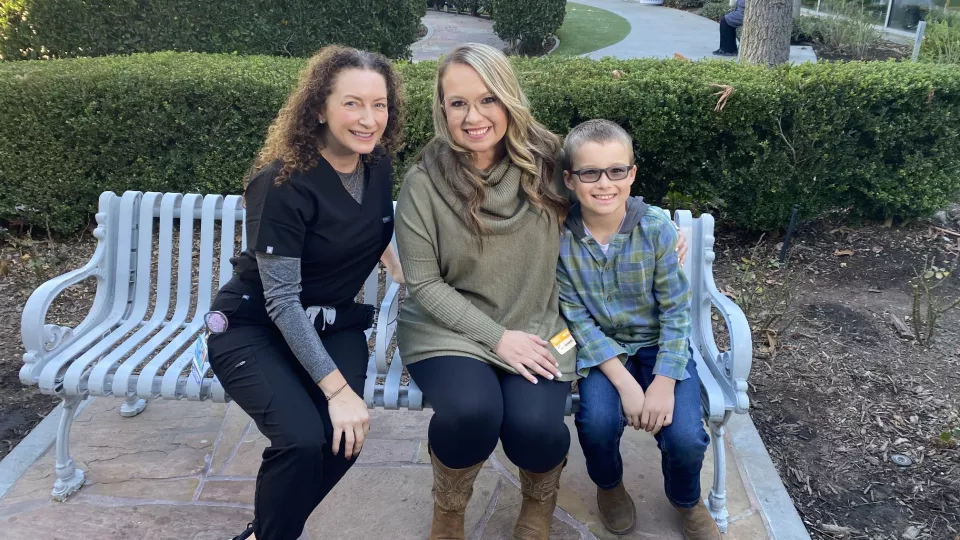 An adult with a light skin tone wearing hospital scrubs and sitting next to an adult and a child with light skin tones and casual clothes, all of whom are smiling.
