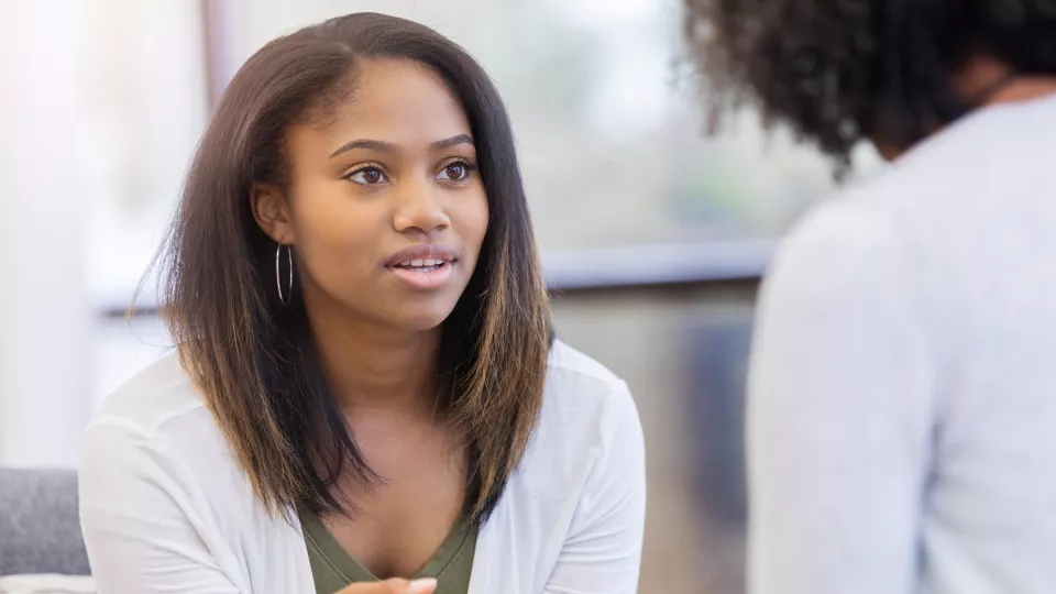 Teen girl with medium skin and shoulder-length dark hair gestures as she talks to a therapist. The therapist’s back is to the camera.