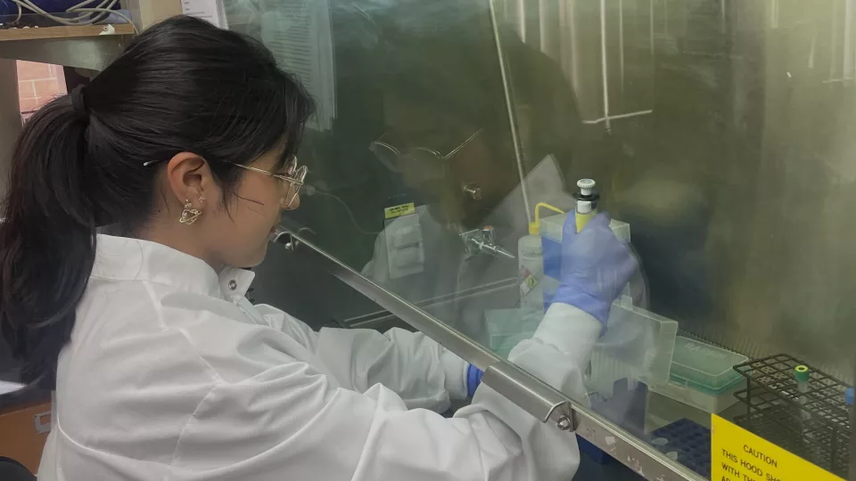 A young adult with a medium skin tone wears a white lab coat, glasses, and gloves and sits in a laboratory, holding various instruments used in medical research.