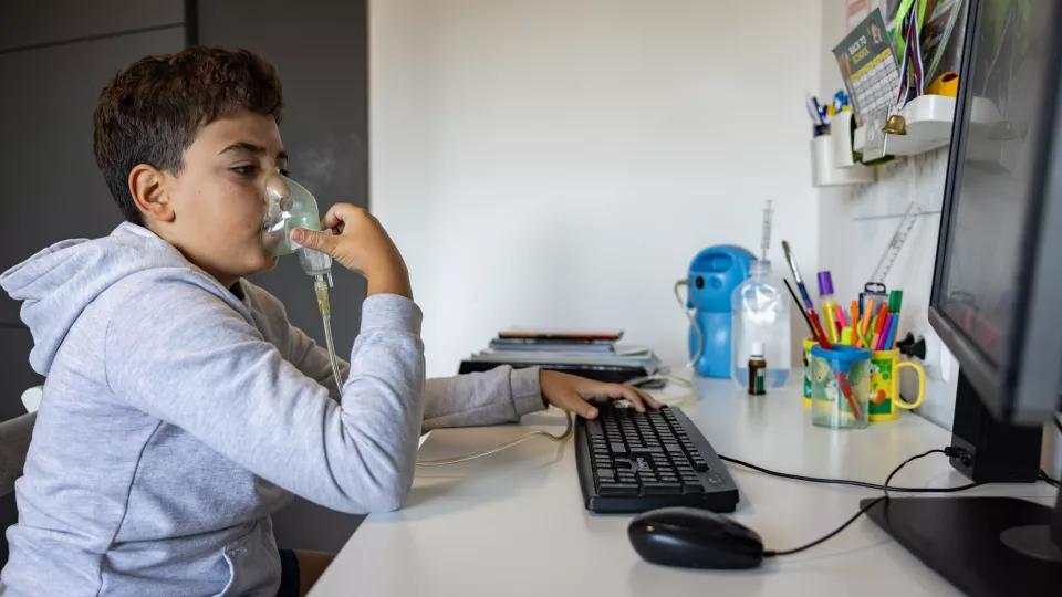 A child with medium skin tone holding an oxygen mask up to his face while sitting at a desk indoors and using a computer.
