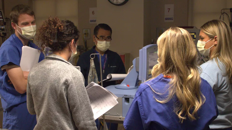 A group of five physicians wearing surgical masks compare notes in a hospital simulation room setting