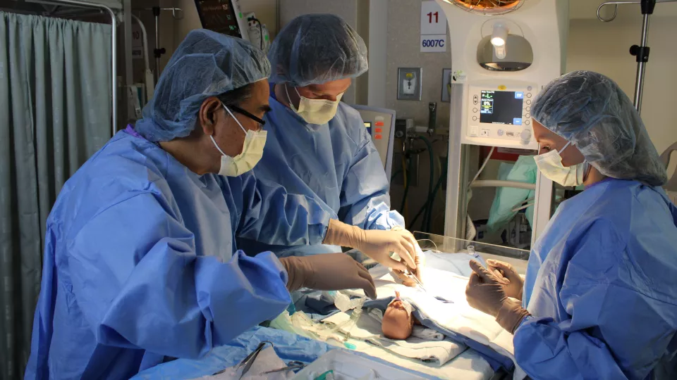 Three surgeons wearing operating gowns, surgical masks and head coverings interact with an infant medical mannequin in a simulated hospital operating room