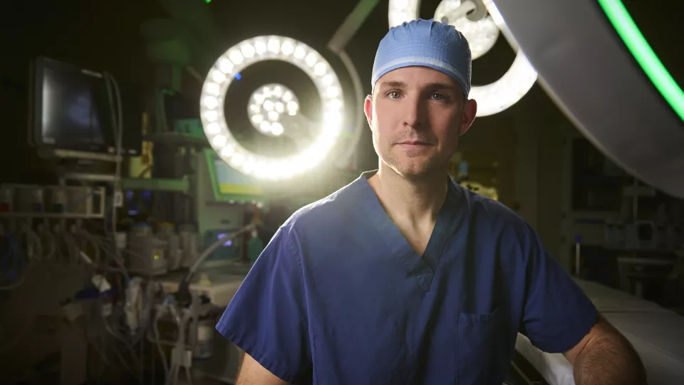 A man with light skin tone wearing dark blue scrubs and a light blue surgical cap gazes at the camera with a serious expression in an operating room. Two circular white bright lights are behind him.