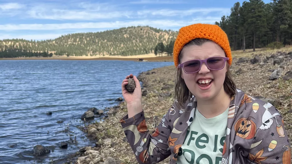 A teenage girl, holding up a small rock, sits by a an alpine lake with mountains behind her.