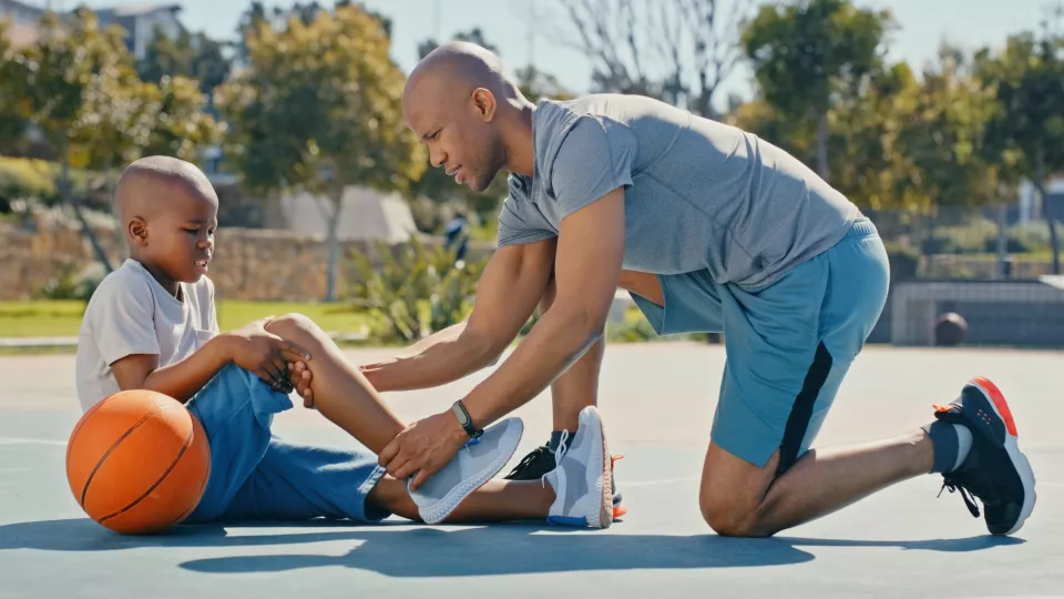 Child with basketball holding right knee in pain while being helped by father