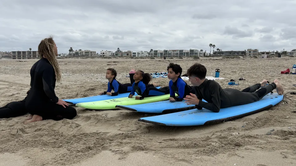 A surf instructor teaches 4 children in wetsuits the "cobra" pose on their boards while they practice on the sand. 
