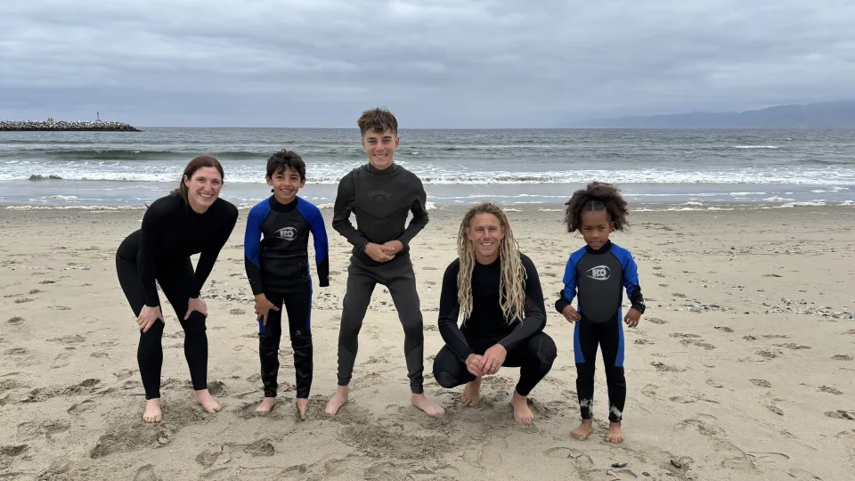 Three kid participants, and two adults pose in wetsuits in front of the ocean. 