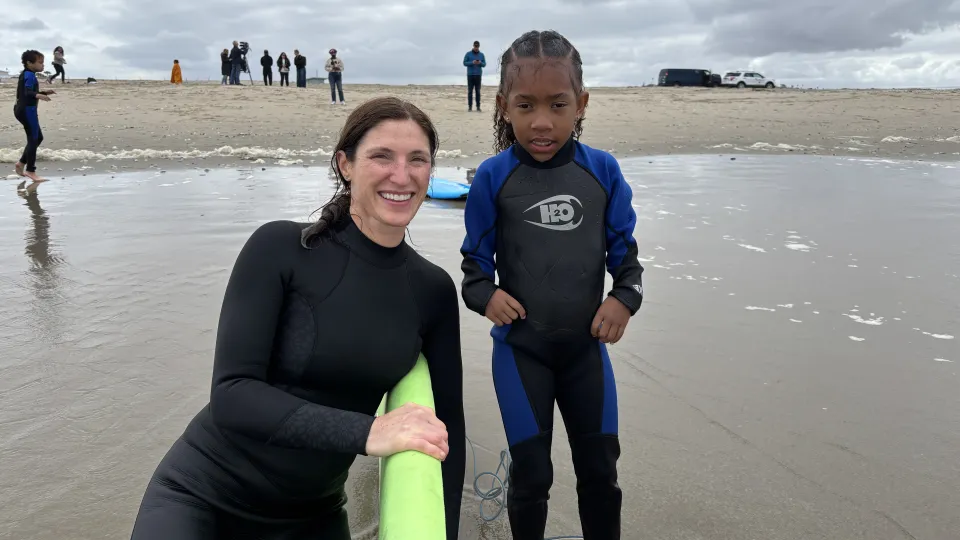 An adult holds a surfboard and poses with a little girl at the beach. Both wear wetsuits. 