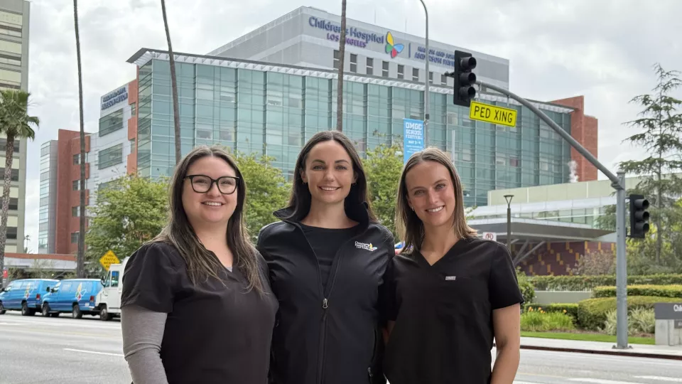 Three nurses wearing black scrubs stand on Sunset Boulevard with Children's Hospital Los Angeles behind them. 
