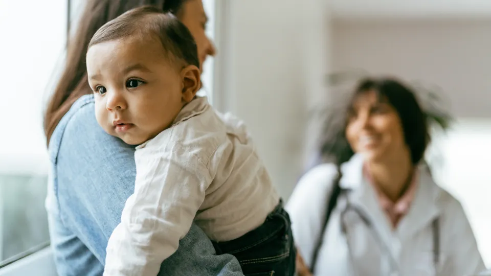 A toddler rests their head on their parent's shoulder as the parent talks with the pediatrician in the background.