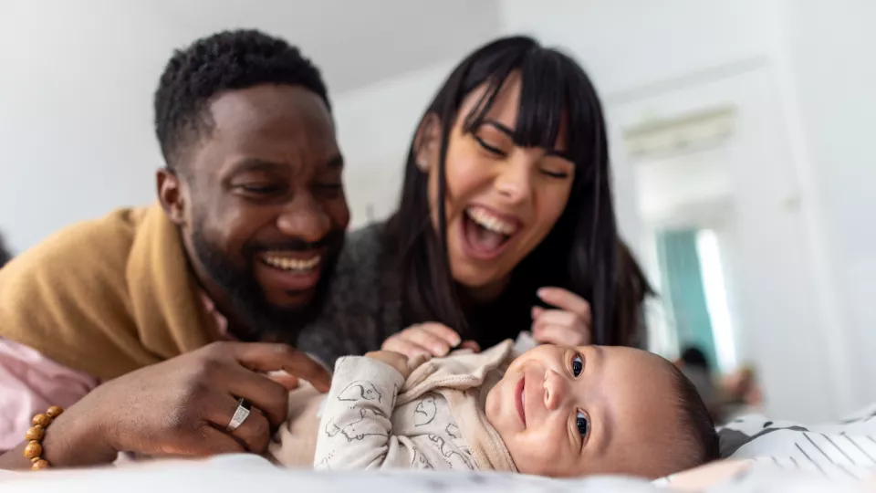 A dark skin toned man and a medium-light skin toned woman smile at a medium-light skin toned baby, also smiling.