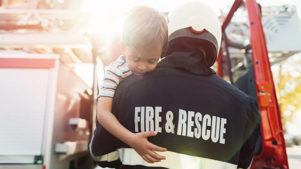 Rear view of a firefighter carrying a young boy over his shoulder toward a red fire truck. The back of the firefighter's jacket says Fire and Rescue.
