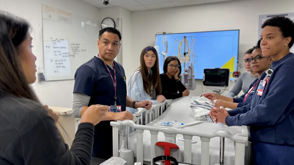 A group of nurses gather around a hospital crib and look toward an instructor.
