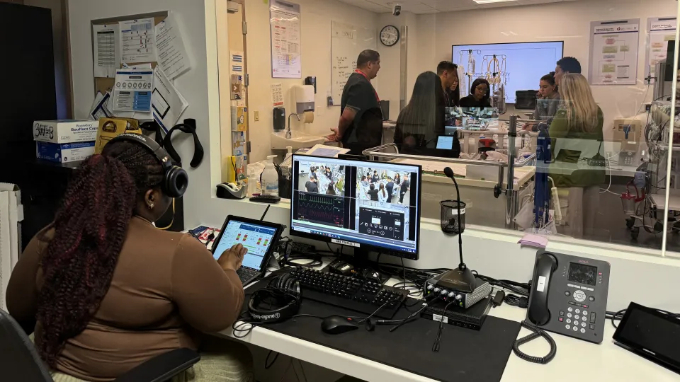 A person sits at a control center which features a tablet, monitor, microphone, and headphones, while looking into a 2-way mirror.