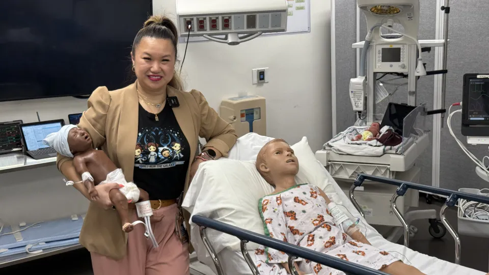A woman stands in a mock hospital room holding a simulation "manikin," surrounded by two other "manikins."