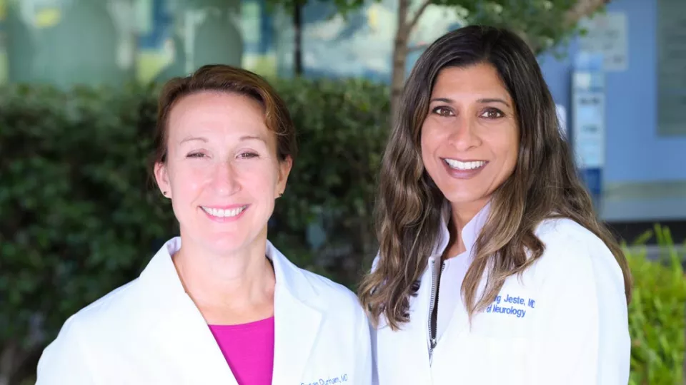 A light skin-toned woman and a medium-dark skin-toned woman, both in lab coats, smile at the camera.