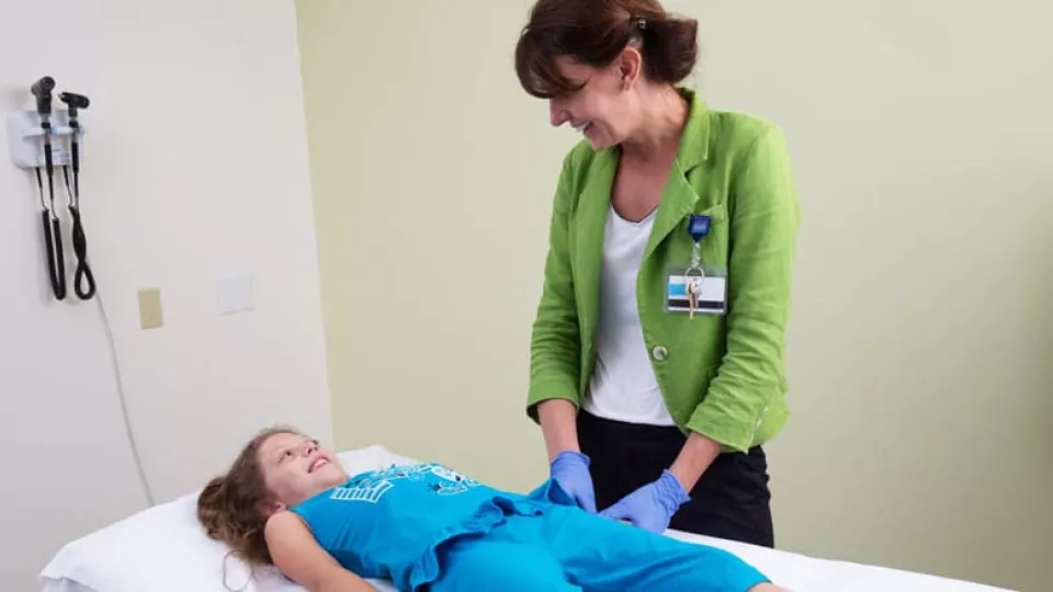 A light skin-toned young girl laying on an exam table and a light skin-toned woman wearing a green jacket and ID badge smile at each other.