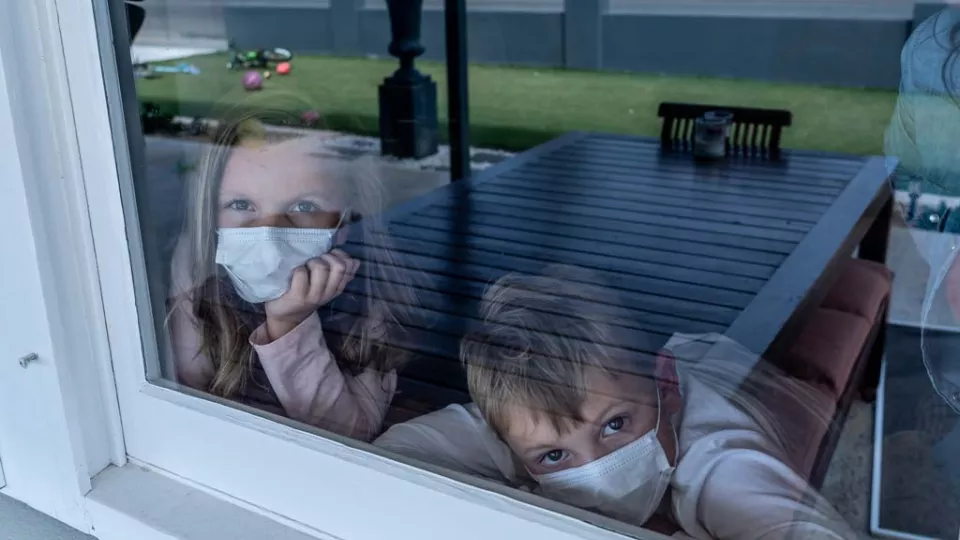 Young girl and boy wearing face masks, looking outside from behind a closed window